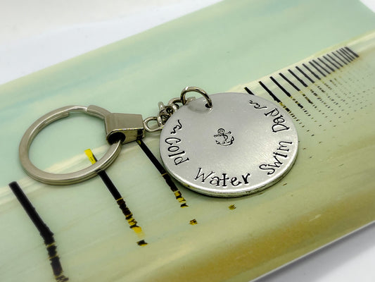 A silver coloured aluminium disc keyring, handstamped with the words Cold Water Swim Dad and an anchor design