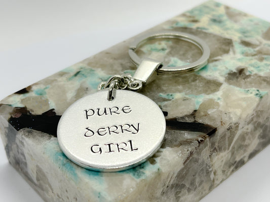 A silver coloured aluminium disc keyring, handstamped with the words Pure Derry Girl