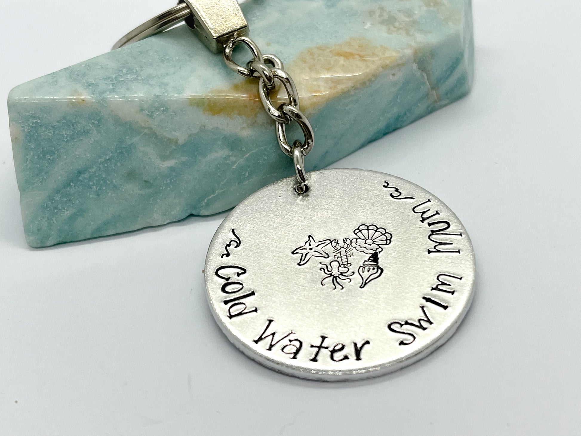 A silver coloured aluminium disc keyring, handstamped with the words Cold Water Swim Mum and ocean designs
