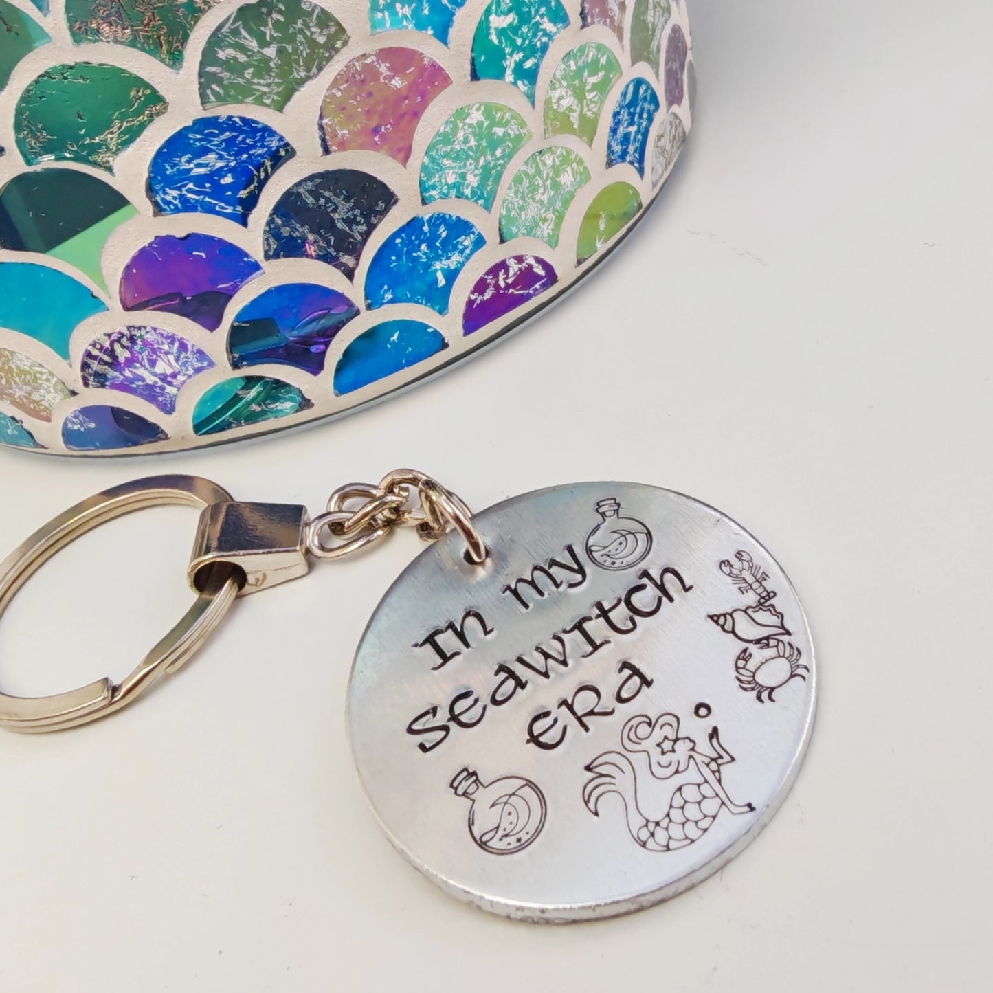A silver coloured aluminium disc keyring, handstamped with the words In My Seawitch Era and a mermaid design