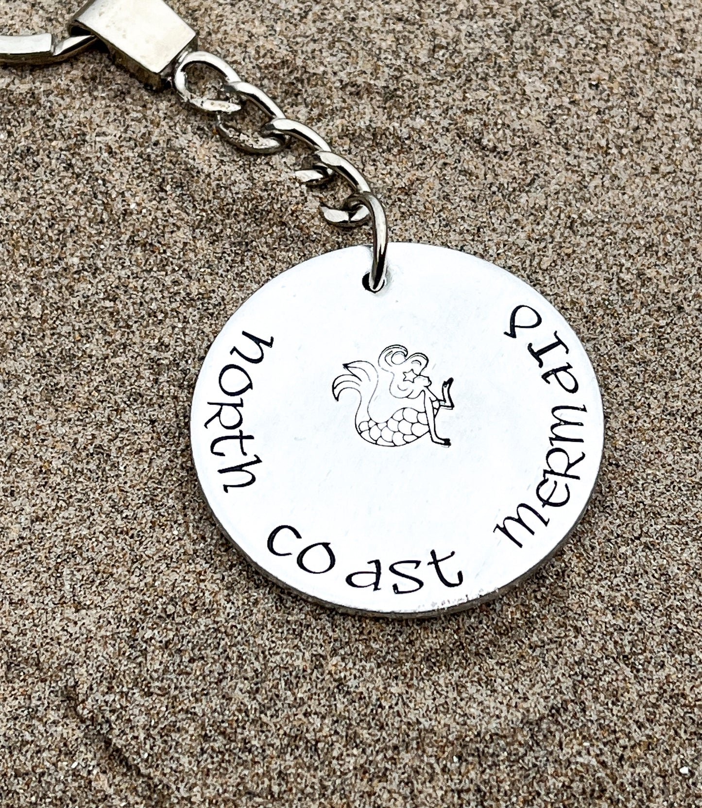 A silver coloured aluminium disc keyring, handstamped with the words North Coast Mermaid and a mermaid design