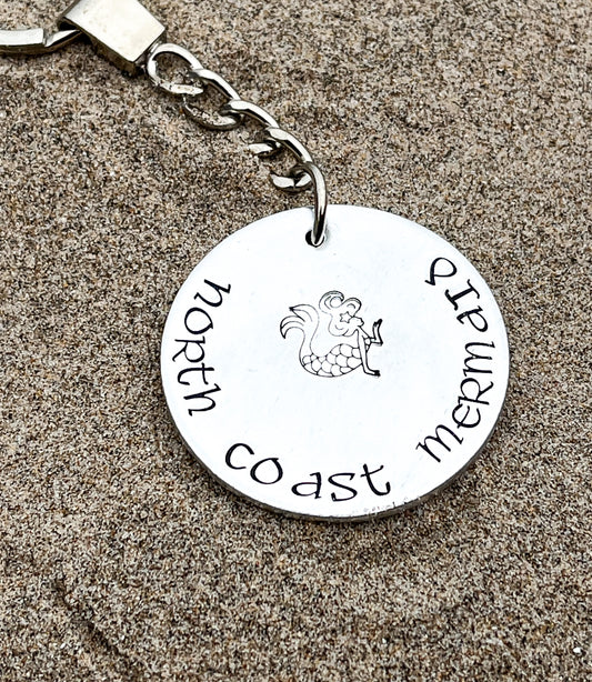 A silver coloured aluminium disc keyring, handstamped with the words North Coast Mermaid and a mermaid design