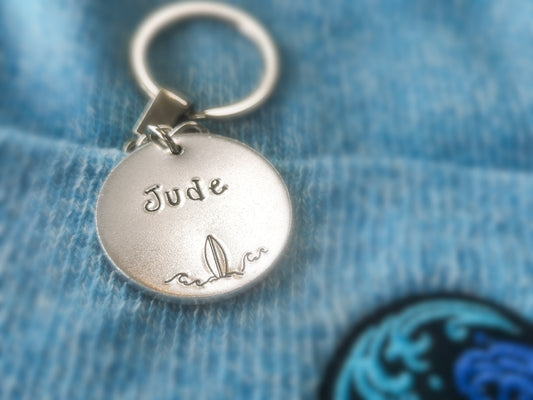 A silver coloured aluminium disc keyring, handstamped with a name and surfboard design